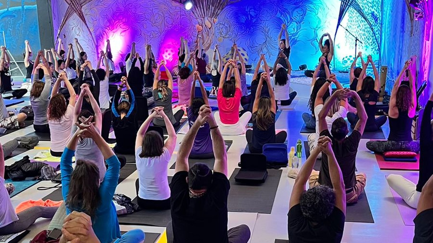 Público sentado em tapetes, de costas, com os braços erguidos durante prática de yoga em sala fechada com luz lilás e azul